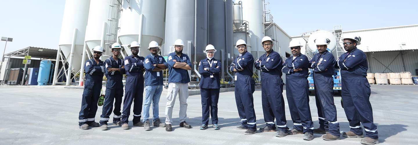 Industrial workers in blue uniforms and safety helmets standing in a line at an industrial facility with large storage tanks in the background